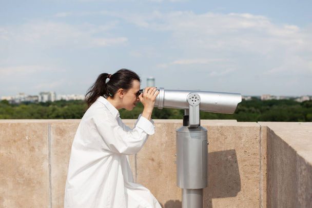 天体望遠鏡を通して見る屋上の建物に立つ白人女性 - 写真・画像