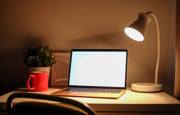 Tietokoneen kannettava tietokone tyhjä näyttö, kahvikuppi ja lamppu valkoisella pöydällä pimeässä huoneessa. - Valokuva, kuva