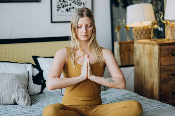 Blonde woman meditates with closed eyes sitting on bed - Foto, afbeelding