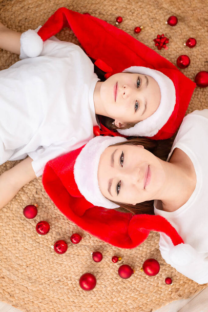 Χαριτωμένες αδελφές σε καπέλα Santa Claus βρίσκονται στο πάτωμα μεταξύ κόκκινα μπάλες Χριστούγεννα στο σπίτι. Στην κορυφή. Χριστούγεννα. Μια εποχή θαυμάτων και εκπλήρωσης επιθυμιών. Καλά Χριστούγεννα.. - Φωτογραφία, εικόνα