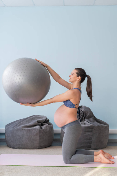 Pregnant european woman performs exercises with fitness ball. Expectant mother is doing yoga in the 3rd trimester. Girl in the classroom in preparation for childbirth. - Foto, Imagen