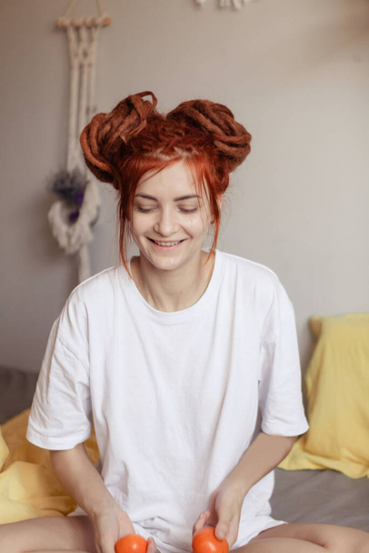 Very pretty redhead girl with dreads He holds tangerines in his hands. A woman in a white T - shirt is sitting in bed . The concept of home recreation during the Christmas holidays - Foto, Imagen