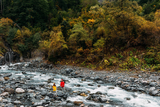 A couple of tourists walking along a mountain river in autumn. Active rest on the nature. An active couple is engaged in Hiking. A young couple is engaged in tracking. Copy space - Foto, imagen