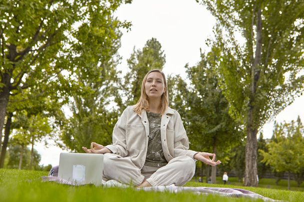 Una mujer se sienta con las piernas cruzadas en la posición de loto Sukhasana y medita en el parque. Mujer atractiva alivia el estrés meditando en el parque - Foto, Imagen