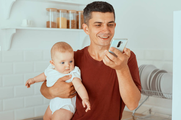 Indoor shot of attractive dark haired father holding infant daughter in hand and using mobile phone for video call, looking at device screen with cute smile. - Foto, afbeelding