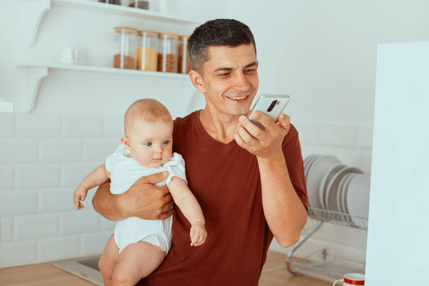 Horizontal shot of smiling handsome man wearing casual style burgundy t shirt standing in kitchen with toddler daughter in hands and watching video on cell phone. - Φωτογραφία, εικόνα