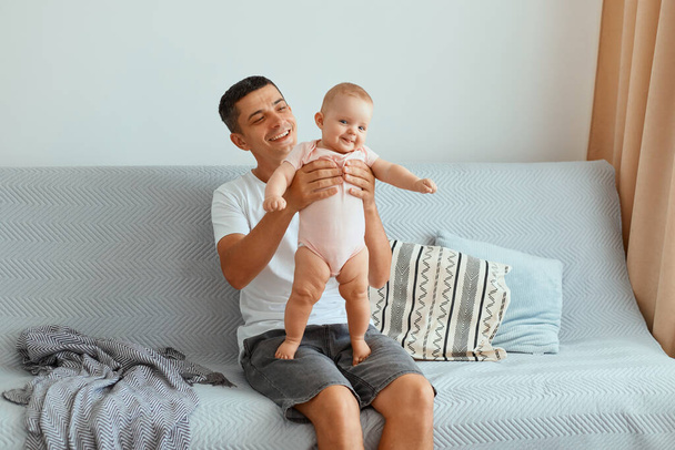 Indoor shot of young adult dark haired man wearing white t shirt and jeans short sitting on cough, happy father holding infant daughter in hands, expressing positive emotions. - Photo, image