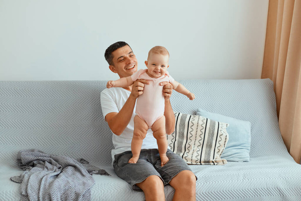 Portrait of Caucasian dark haired man wearing white t shirt and jeans short sitting on sofa and holding happy smiling toddler daughter, happy father spending time with his kid. - Photo, image