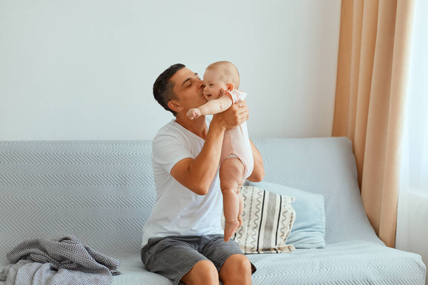 Image of handsome brunette father wearing white t shirt and jeans short sitting on sofa with his charming daughter and kissing her, expressing love and care. - Zdjęcie, obraz