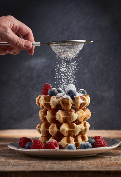 Woman's hand sprinkling powder sugar on a stack of waffles with fresh berries on them - Foto, afbeelding