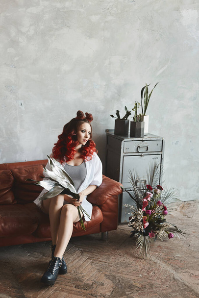 Kırmızı saçlı, tulumlu ve düğmesiz gömlekli muhteşem bir genç kadın çatı katındaki koltukta oturuyor. - Fotoğraf, Görsel