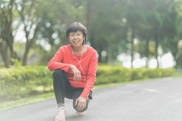 シニアアジアの女性ランナーの笑顔はフィットネスモチベーションのためにイヤホンで音楽を聴く進行状況を追跡するモバイルスマートフォンアプリを使用しています。公園で走る準備をしているアジアの女性 - 写真・画像