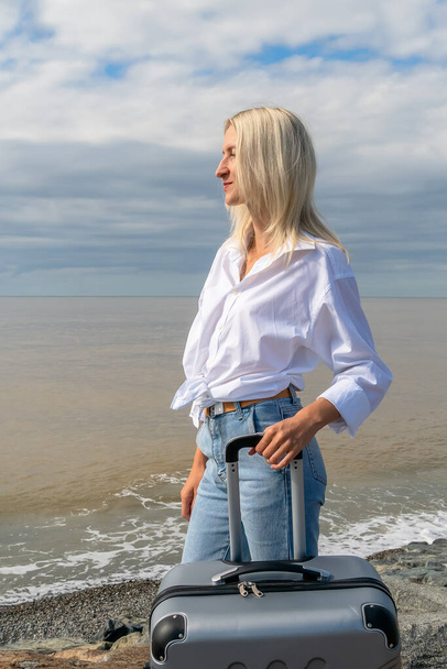 Nuori blondi nainen hymy farkut ja paita harmaa matkalaukku seisoo meren rannalla pilvisellä säällä. Pystysuuntainen - Valokuva, kuva