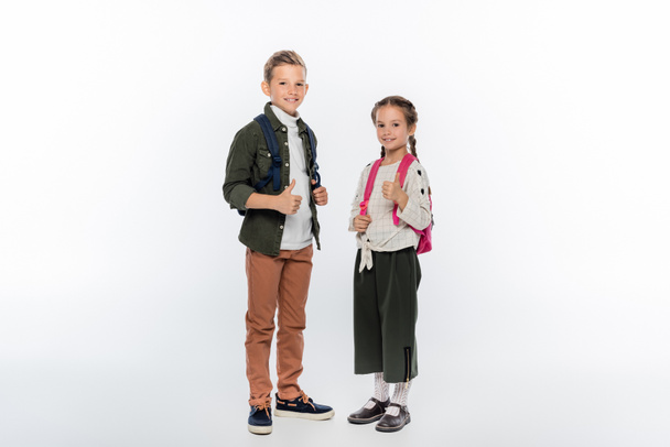 cheerful schoolkids standing with backpacks and showing thumbs up on white  - Foto, afbeelding