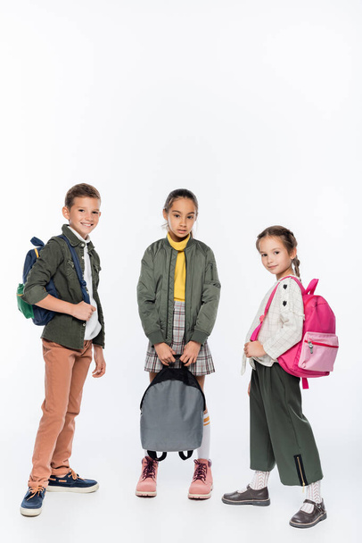 cheerful schoolboy and smiling schoolgirls standing with backpacks on white  - Foto, Imagen