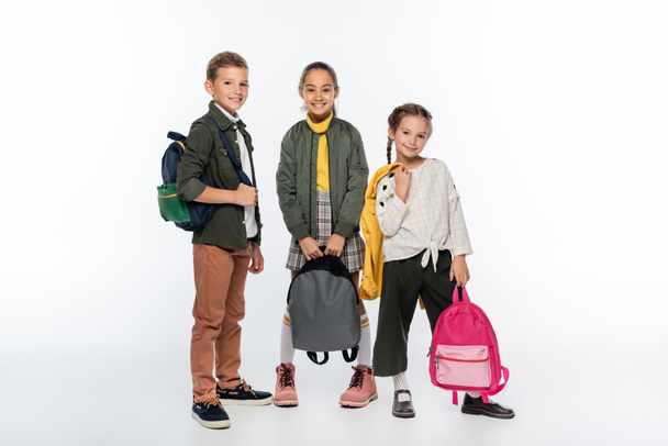 cheerful schoolboy and positive schoolgirls standing with backpacks on white  - Fotografie, Obrázek