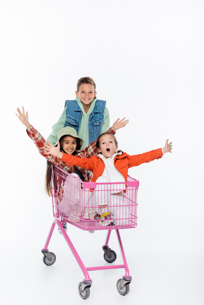 boldog fiú áll mögötte csodálkozó lányok bevásárlókocsiban elszigetelt fehér  - Fotó, kép