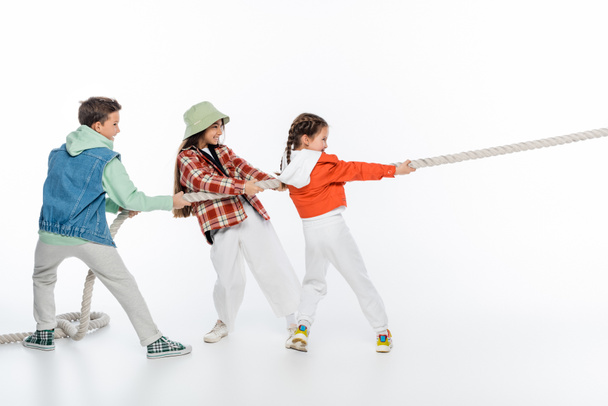 visão lateral de crianças pré-adolescentes felizes puxando corda enquanto joga rebocador de jogo de guerra no branco - Foto, Imagem