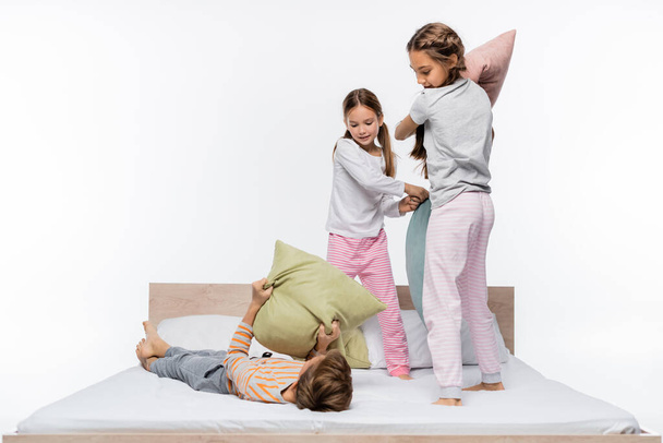 szczęśliwy dziewczyny w piżamie mając walki na poduszki stojąc obok chłopca na łóżku odizolowany na biały - Zdjęcie, obraz