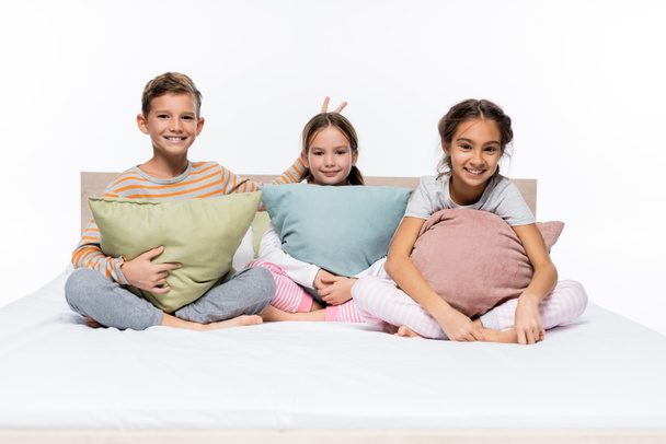 happy boy showing bunny ears with fingers near girl and friend with pillows isolated on white - Photo, image