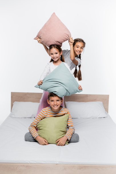 playful girls in pajamas having pillow fight with boy while standing isolated on white - Fotografie, Obrázek
