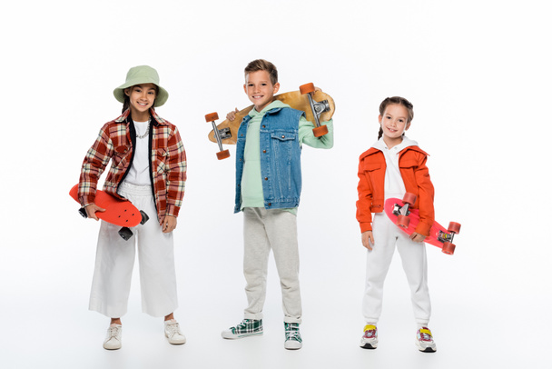full length of trendy children smiling while holding longboard and penny boards on white - Foto, Imagem