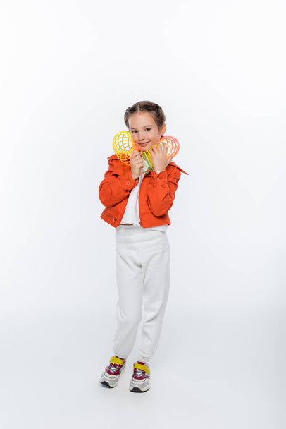full length of happy kid in orange jacket playing with rainbow slinky on white - Фото, изображение
