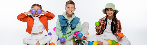 cheerful preteen kids sitting near friend covering eyes with colorful balls on white, banner - Photo, Image