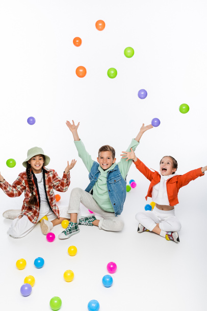 alegre preadolescente niños sentado y lanzando bolas de colores en el aire en blanco  - Foto, imagen