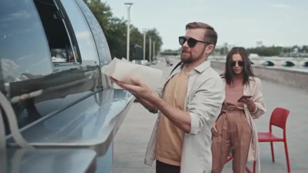 Μεσαία βολή του νέου άνδρα και της γυναίκας που αγοράζουν χοτ ντογκ από φορτηγό τροφίμων στέκεται έξω την ηλιόλουστη μέρα, πληρώνουν με smartphone - Πλάνα, βίντεο