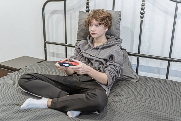 teenage girl short brown curly hair, dressed in unkempt home clothes, sits unmade bed and plays computer console. Passion for computer games, addiction, hobbies. To spend time in isolation, waste of time. teenager's room is colorless and impersonal - Photo, Image