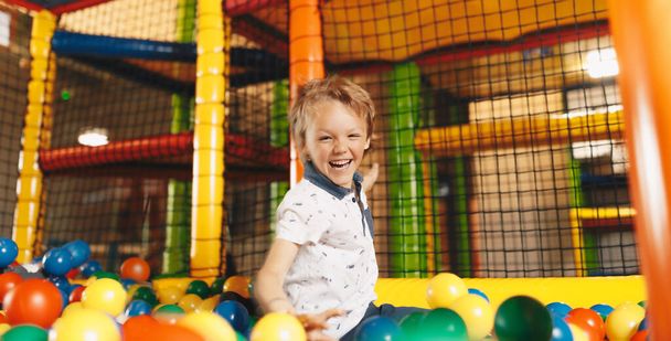 Happy Boy skaczący do basenu z piłkami w parku rozrywki. Dzieciak bawiący się w przedszkolu. Chłopiec bawiący się w kolorowy basen wypełniony plastikowymi kulkami - Zdjęcie, obraz
