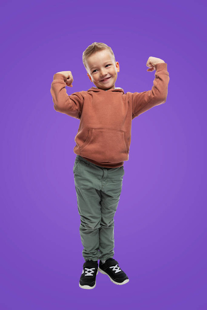 A 5-year-old boy stands and shows muscles in his arms. A child in jeans and a brown sweater. Sports and children's health. Purple background. Full height. Vertical. - Фото, изображение