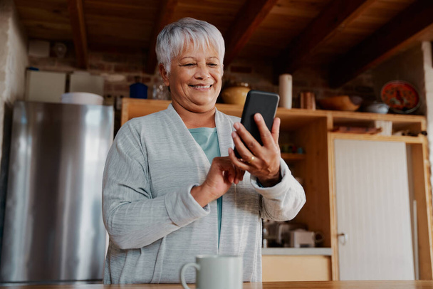 Multi-cultural elderly female reading daily news on smartphone while standing in modern kitchen. - Φωτογραφία, εικόνα