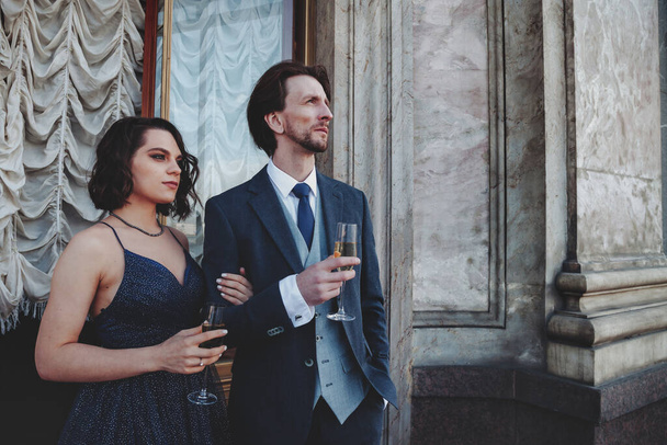 Couple, man and woman, with champagne on balcony of historical palace in evening dresses, suit and dress. An elderly couple in love spends weekend. Concept of romantic dates and surprises. Copy space - 写真・画像