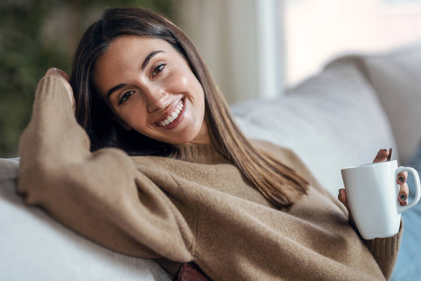 自宅のリビングルームでソファに座っているカメラを見ながらコーヒーを飲みながら美しい若い女性のショット. - 写真・画像
