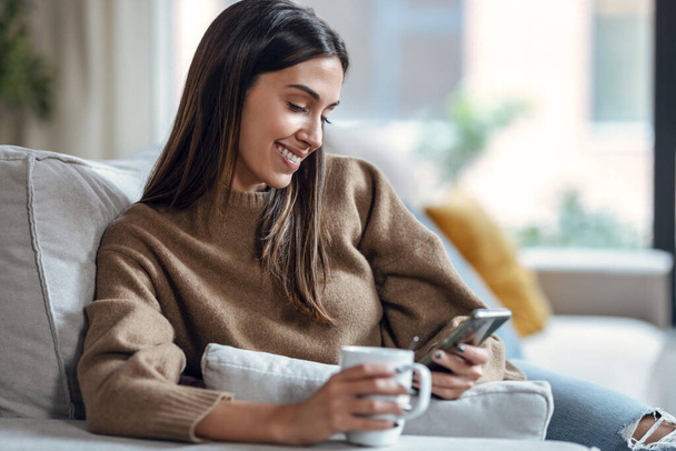 Shot of smiling young woman using her mobile phone while drinking coffee sitting on sofa at home. - Foto, Imagen