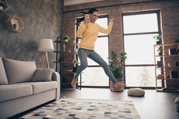 Photo of funny sweet young lady wear yellow pullover rising fist holding modern gadget jumping indoors room home - Fotoğraf, Görsel