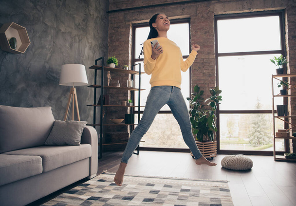 Photo of lucky funny young lady wear yellow pullover rising fist holding modern gadget jumping indoors room home - 写真・画像