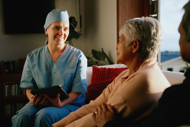 Medical professional smiling at elderly multi-ethnic couple. Home consultation in modern living room. - Foto, imagen
