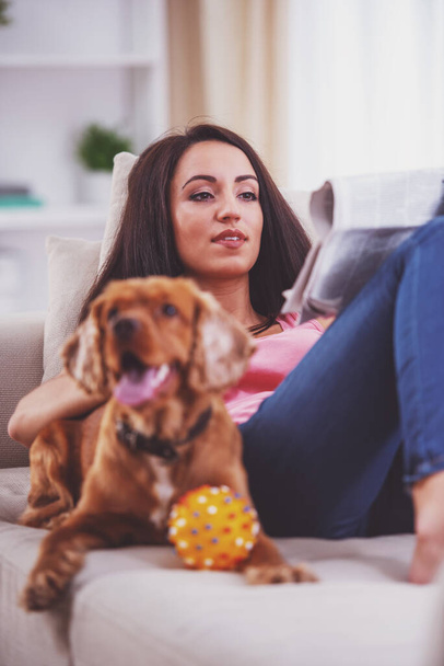 Bild einer jungen Frau liest Zeitung, während ihr Hund neben ihr auf dem Sofa liegt. - Foto, Bild
