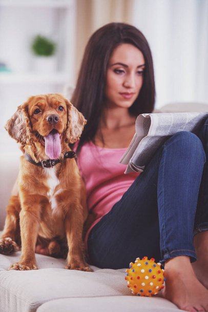 彼女の犬は彼女の近くのソファに座っている間に若い女性の写真が新聞を読んでいます。. - 写真・画像