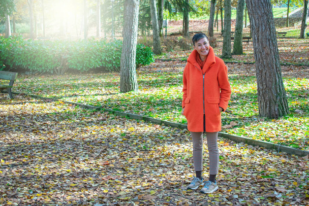 елегантна жінка в помаранчевому пальто позує посміхаючись в парку
 - Фото, зображення