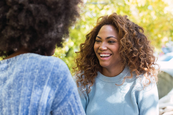 Портрет африканской американки с идеальными белыми зубами, улыбающейся подруге. Афро-черные друзья с вьющимися волосами и голубым свитером, улыбающиеся во время разговора. Концепция отношений - Фото, изображение