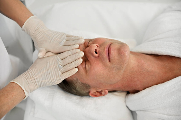 Close-up de um homem europeu maduro relaxante durante a massagem de levantamento de rosto, enquanto um esteticista massagear a testa na clínica de bem-estar spa. Cosmetologia masculina e conceito de cuidados com a pele - Foto, Imagem