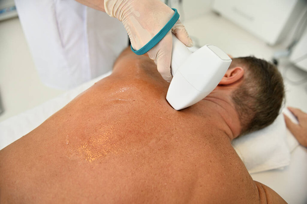 Radiogolven heffen in de spa kliniek. Close-up van de rug van wellness spa mannelijke klant die professionele lichaamsbehandeling met moderne medische apparatuur- Apparatuur voor ultrasone cavitatie van het lichaam - Foto, afbeelding