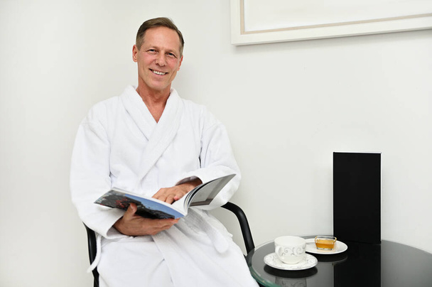 Handsome charming mature European man in white terry bathrobe sitting on armchair with a magazine in his hands, smiles toothy smile looking at camera, relaxing at luxurious wellness spa centre - 写真・画像