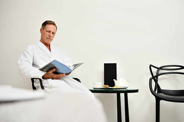 Schöner, attraktiver, reifer europäischer Mann im weißen Frottee-Bademantel sitzt auf einem Sessel und liest ein Magazin und entspannt sich in einem privaten Zimmer im luxuriösen Wellness-Center - Foto, Bild
