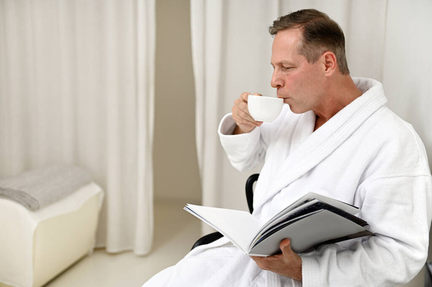 Beau et imposant homme européen de 50 ans vêtu de peignoir éponge blanc, buvant une tisane saine et lisant un magazine assis sur un fauteuil dans une salle privée du centre de spa de luxe - Photo, image