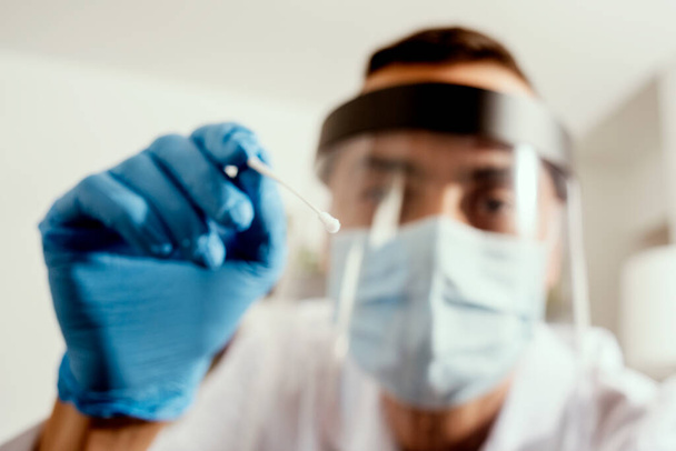 closeup of a male healthcare worker, wearing a white coat, surgical mask, face shield and blue surgical gloves, about to take a nasopharyngeal culture with a swab - Photo, Image